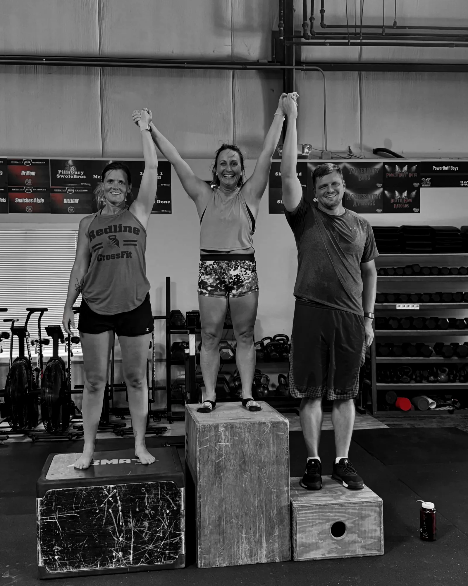 Three Redline members on a plyo-box podium with arms raised together after a workout at the Waukee gym